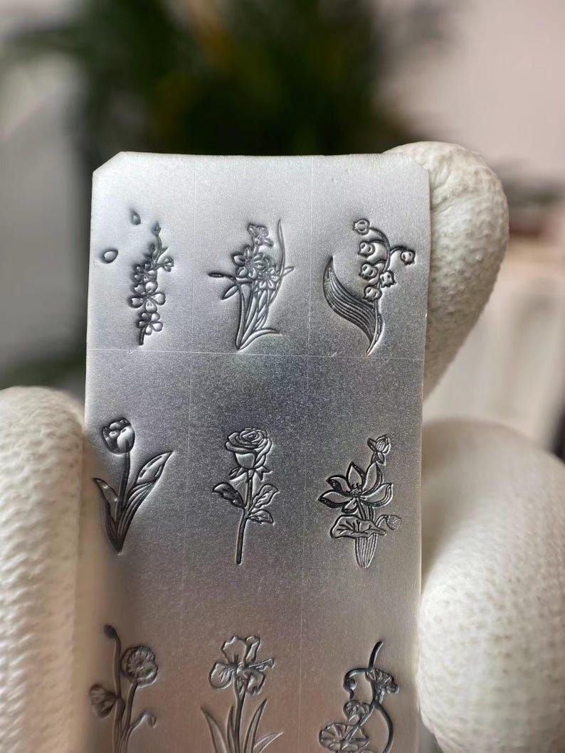 Pu&ograve; includere: Una piastra di metallo argentata con nove diversi disegni di fiori incisi su di essa. I disegni includono mughetto, una rosa, un tulipano, un iris e altri motivi floreali.