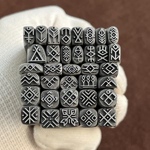 May include: A collection of metal stamps with various geometric and abstract designs. The stamps are arranged in a grid, showcasing a variety of shapes and patterns. The stamps are held by a gloved hand.