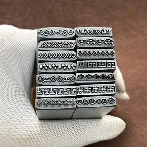 May include: A set of 14 metal stamps with different decorative border designs. The stamps are arranged in a stack and are held in a gloved hand.