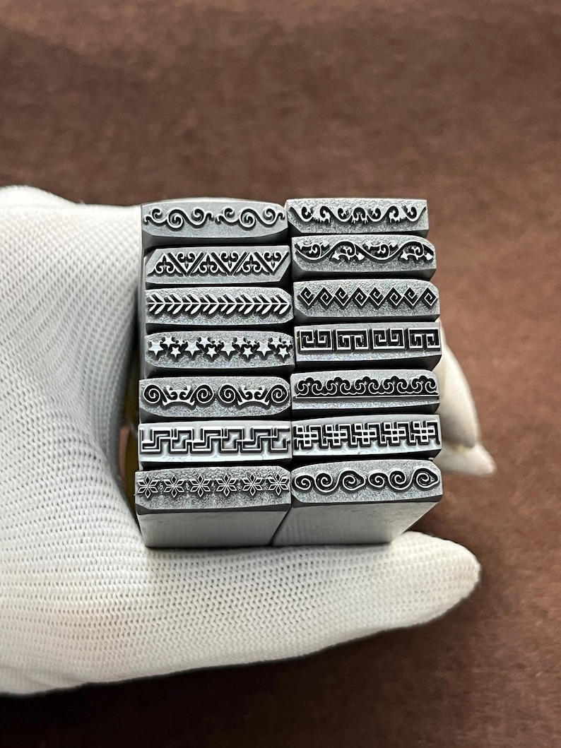 May include: A set of 14 metal stamps with different decorative border designs. The stamps are arranged in a stack and are held in a gloved hand.