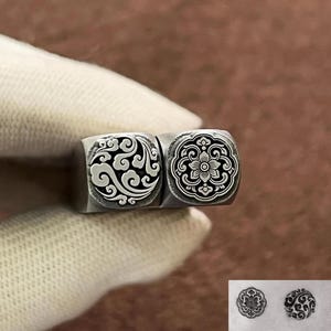 Könnte beinhalten: Zwei Metallstempel mit aufwendigen Designs. Einer zeigt ein wirbelndes Wolkenmuster, der andere ein Blumenmotiv. Die Stempel werden von einer Hand gehalten, was die detaillierte Handwerkskunst zeigt. Die Designs sind in Schwarz und Weiß.
