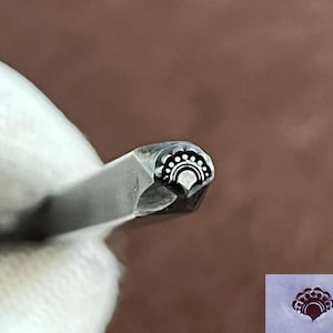 Puede incluir: Primer plano de una herramienta de sello metálico con un diseño decorativo en forma de abanico. El sello presenta una serie de pequeños puntos blancos y una línea curva, creando un patrón detallado. La herramienta es sostenida por una mano enguantada, con un fondo marrón.