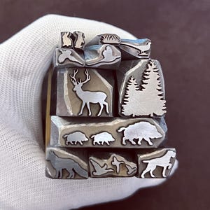 May include: A set of metal stamps featuring various woodland animals and plants, including a deer, boar, birds, rabbits, and trees. The stamps are arranged in a stack and are held in a gloved hand.