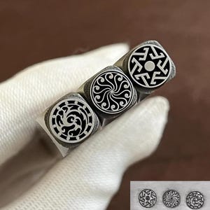 Pode incluir: Três ferramentas de estampagem de metal com hastes quadradas, cada uma apresentando um design circular gravado exclusivo na face. Os designs incluem um padrão geométrico, um motivo de espiral giratória e um padrão complexo, exibidos com impressões.