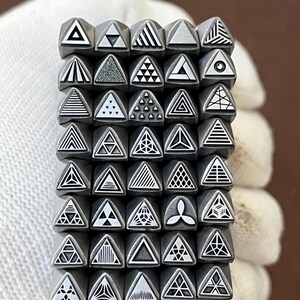 Puede incluir: Un conjunto de sellos metálicos con cabezas triangulares, cada uno con un diseño geométrico único. Los sellos están dispuestos en una cuadrícula, mostrando varios patrones como líneas, puntos y formas dentro de los triángulos. Los sellos son sostenidos por una mano enguantada.