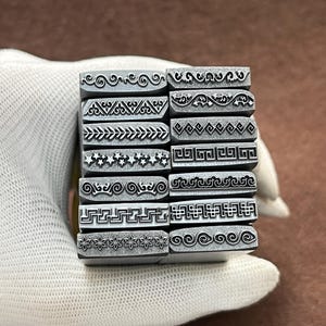 May include: A set of 12 metal stamps with different decorative border designs. The stamps are arranged in a stack and are held in a gloved hand.