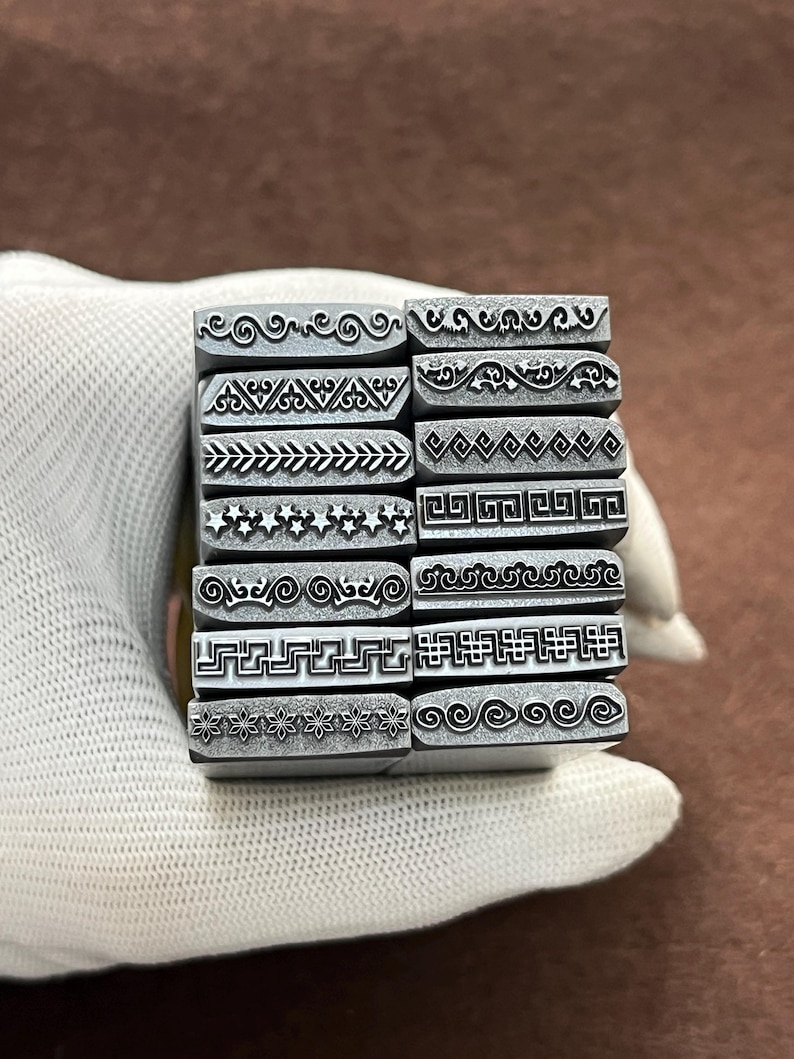 May include: A set of 12 metal stamps with different decorative border designs. The stamps are arranged in a stack and are held in a gloved hand.