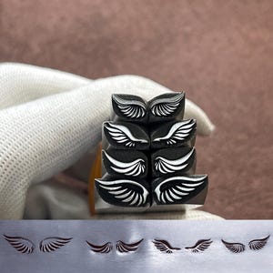 Puede incluir: Un conjunto de sellos de metal negro con un diseño de alas. Los sellos están apilados, con las impresiones de las alas visibles en una superficie metálica debajo. Las alas están detalladas con líneas blancas, para una estética limpia.