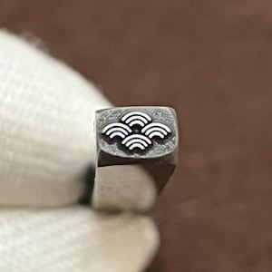 Puede incluir: Un pequeño sello de metal con cara cuadrada que presenta un diseño empotrado de cuatro arcos semicirculares concéntricos y superpuestos dispuestos en grupo. El sello tiene un acabado metálico oscuro y texturizado en su cara.