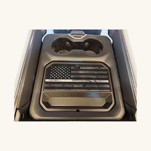 Può includere: Un vassoio organizer per console centrale auto di colore nero con un design a bandiera americana testurizzato e una sottile linea orizzontale. Il vassoio include uno scomparto rettangolare ed è posizionato in una console con due portabicchieri.