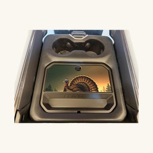 May include: A black automotive centre console insert featuring a graphic of a wild turkey with its tail feathers fanned out against a sunset forest background. The insert is positioned in front of two circular cup holders in a vehicle interior.