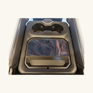 May include: A black automotive center console insert featuring two circular cup holders and a rectangular storage tray. The tray is covered with a dark surface displaying a red and blue topographic map pattern with intricate contour lines.
