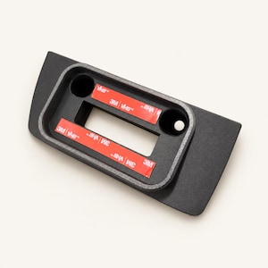 May include: A matte black rectangular mounting bracket featuring a central rectangular cutout and two circular holes on the sides. Two strips of red 3M VHB adhesive tape are applied horizontally across the surface for secure installation.