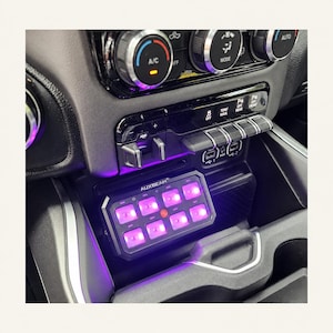 May include: An Auxbeam switch panel with eight rectangular buttons illuminated in bright purple, mounted in a vehicle centre console below the climate control dials, featuring a black housing and a central circular power button icon.