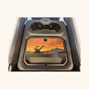 Op de afbeelding: Een zwarte middenconsole-inzet voor in de auto met een rechthoekig paneel voorzien van een afbeelding van een eland in een berglandschap bij zonsondergang. De inzet bevat twee ronde bekerhouders en een opbergvak onder het grafische paneel.