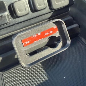 Puede incluir: Accesorio de coche de plástico negro con marco rectangular y recorte circular. El accesorio tiene una tira adhesiva roja y blanca en la parte posterior, diseñado para encajar en la consola de un coche.