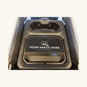 May include: A black automotive center console insert featuring two circular cup holders and a rectangular recessed area. The center section displays a graphic with an image icon and the white text "YOUR IMAGE HERE Any photo, logo or artwork."