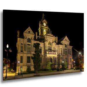 May include: A historic stone courthouse building illuminated at night, featuring a prominent central clock tower, multiple arched windows, and decorative bunting on the balcony. The structure is set against a dark sky with street lamps.