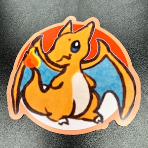 Könnte beinhalten: Ein Aufkleber mit einem Cartoon-Charizard. Der orangefarbene und weiße Aufkleber zeigt ein Cartoon-Bild von Charizard mit blauen Flügeln, einer Flamme am Schwanz und einem roten und orangefarbenen Hintergrund.