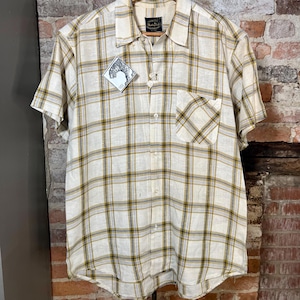 May include: Short-sleeved button-down shirt in a cream and olive green plaid pattern. The shirt has a single chest pocket and a collar. A tag with a bear graphic is attached. The shirt is hanging on a wooden hanger that reads "HOTELS STATLER".