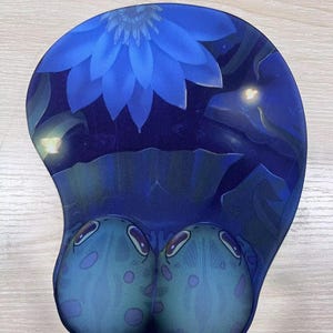 May include: Blue ergonomic mouse pad with a floral design and a frog-like shape. The pad features a large blue flower at the top and two cartoonish frog-like shapes at the bottom. The design is set against a dark blue background.