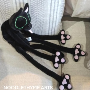 Könnte beinhalten: Ein schwarzes Plüschkätzchen mit langen, flexiblen Gliedmaßen und großen, grün umrandeten Augen. Die Pfoten der Katze sind rosa mit schwarzen Details. Der Text "NOODLETHYME ARTS" ist unten im Bild zu sehen.