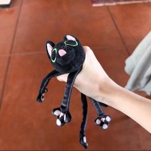 May include: A black plush cat toy with long, dangling limbs and a tail. The cat features embroidered green eyes, a small pink nose, and light pink paw pads on its feet. The plush material is soft and dark, with a distinct head and four limbs.