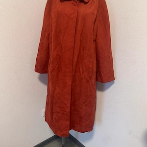 May include: A terracotta-coloured coat with a button fastening and a wide collar. The coat is displayed on a mannequin and appears to be made of a soft, suede-like material. The coat has long sleeves and a classic design.