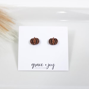 Puede incluir: Un par de pequeños pendientes de botón en forma de calabaza con un acabado de purpurina de cobre. Los pendientes se muestran en una tarjeta blanca con el texto "grace + joy collective".