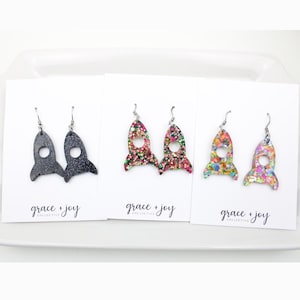 Può includere: Tre paia di orecchini a forma di razzo su biglietti bianchi. Gli orecchini sono neri, glitter rosa e verdi e glitter multicolore. Ogni biglietto ha il testo "grace + joy collective" in scrittura nera.