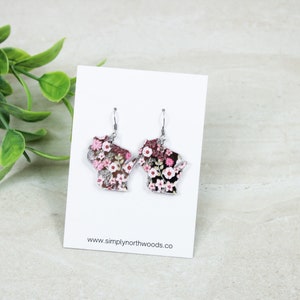 May include: A pair of silver earrings shaped like the state of Wisconsin, with a floral print featuring pink and white flowers on a black background. The earrings are hanging from a white card with the text "www.simplynorthwoods.co".