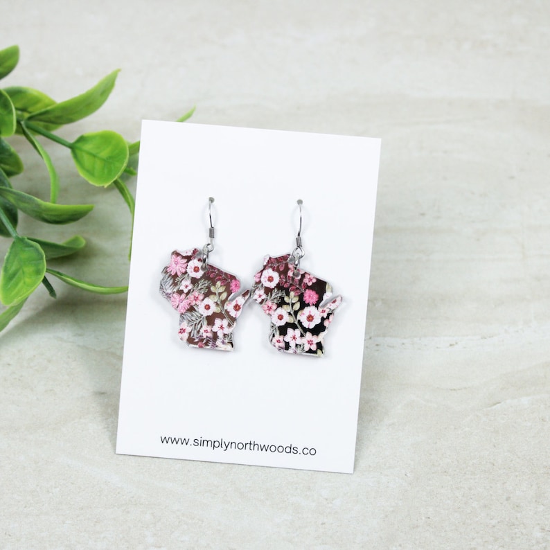 May include: A pair of silver earrings shaped like the state of Wisconsin, with a floral print featuring pink and white flowers on a black background. The earrings are hanging from a white card with the text "www.simplynorthwoods.co".
