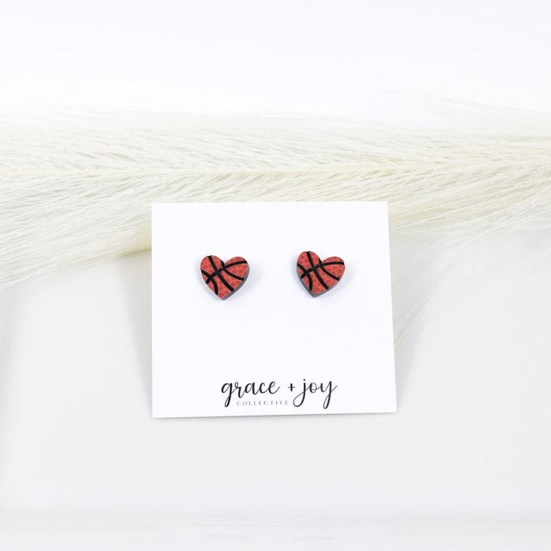 Peut inclure: Une paire de boucles d'oreilles &agrave; clous en forme de c&oelig;ur avec un motif de ballon de basket orange textur&eacute; et des lignes noires. Les boucles d'oreilles sont fix&eacute;es sur une carte de pr&eacute;sentation blanche avec le texte grace + joy collective.