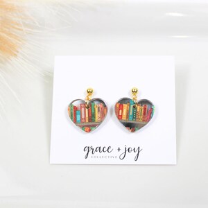 Peut inclure: Une paire de boucles d'oreilles en forme de c&oelig;ur avec un crochet en m&eacute;tal dor&eacute;. Les boucles d'oreilles pr&eacute;sentent un fond transparent avec une image color&eacute;e d'une biblioth&egrave;que remplie de livres.