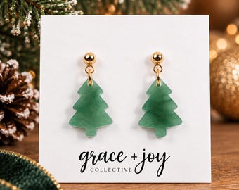 May include: A pair of dangle earrings featuring green marbled acrylic Christmas tree charms suspended from gold-tone ball studs. The earrings are displayed on a white card printed with the text grace + joy COLLECTIVE in black script.