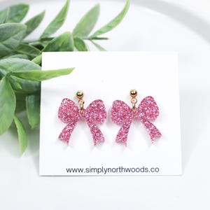 May include: Pink glitter bow earrings with gold posts. The earrings are on a white card with the website www.simplynorthwoods.co printed on it.