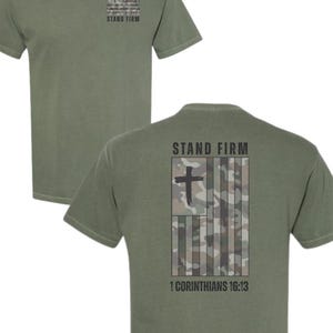Könnte beinhalten: Olivgrünes T-Shirt mit einem Tarnflaggen-Design und den Worten "STAND FIRM" und "1 CORINTHIANS 16:13". Das Shirt hat einen Rundhalsausschnitt und kurze Ärmel. Das Design beinhaltet ein Kreuz.