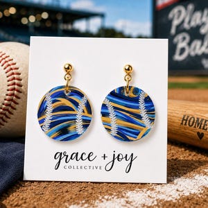 May include: A pair of circular dangle earrings featuring a blue and gold abstract swirl pattern with white baseball stitch details. The earrings are attached to a white display card with the text grace + joy collective printed in black.