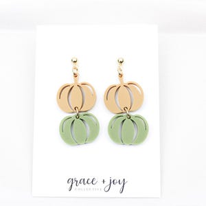 May include: A pair of pumpkin-shaped earrings. Each earring features a light tan pumpkin shape above a green pumpkin shape. The earrings are suspended from gold-colored hooks. The words "grace + joy" are printed in cursive on the bottom of the white card.