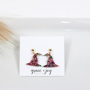 May include: A pair of pink and green glitter witch hat earrings with gold posts. The earrings are on a white card with the text "grace + joy collective".