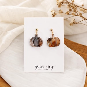 May include: A pair of dangle earrings featuring pumpkin-shaped charms made of brown and tan marbled acrylic. The charms hang from small gold-toned metal posts. The earrings are attached to a white display card with the text grace + joy collective.