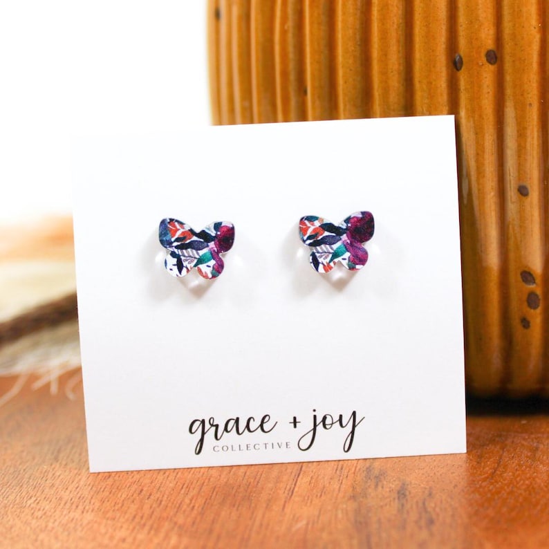 May include: A pair of butterfly-shaped earrings with a floral print in shades of purple, red, green, and white. The earrings are displayed on a white card with the text "grace + joy collective".