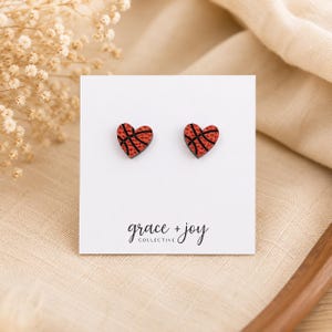 Peut inclure: Une paire de boucles d'oreilles en forme de c&oelig;ur avec un motif de ballon de basket orange textur&eacute; et des lignes noires. Les boucles d'oreilles sont fix&eacute;es sur une carte de pr&eacute;sentation blanche avec le texte grace + joy COLLECTIVE en noir.