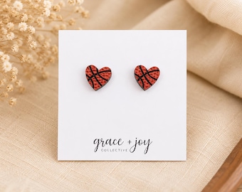 May include: A pair of heart-shaped stud earrings featuring a textured orange basketball pattern with black lines. The earrings are attached to a white display card with the text grace + joy COLLECTIVE printed in black at the bottom center.