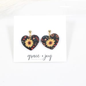 May include: Heart-shaped earrings with a sunflower design. The earrings feature a black background with yellow sunflowers and red accents. The earrings are suspended from gold-colored posts. The words "grace + joy collective" are printed below the earrings.