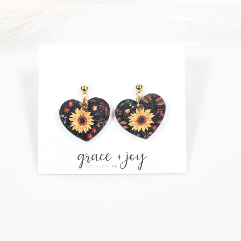 May include: Heart-shaped earrings with a sunflower design. The earrings feature a black background with yellow sunflowers and red accents. The earrings are suspended from gold-colored posts. The words "grace + joy collective" are printed below the earrings.