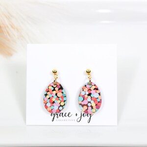 May include: A pair of gold stud earrings with oval-shaped, colorful confetti-patterned dangles. The earrings are on a white background with the text "grace + joy collective".