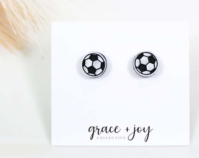 May include: A pair of small, white stud earrings with a black soccer ball design. The earrings are displayed on a white card with the text "grace + joy collective".