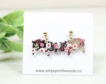 May include: A pair of gold pig earrings with a floral print. The pigs are facing each other and the print is a mix of pink, white, and black flowers.