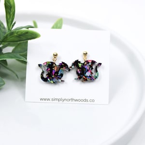 May include: A pair of iridescent black dragon earrings with gold posts. The dragon design is filled with colorful glitter.